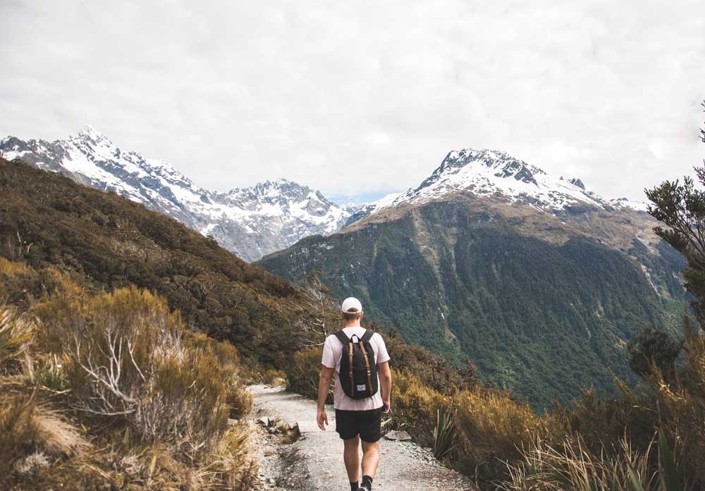 fotografía de un chico de espaldas con una mochila y una gorra andando entre montañas 